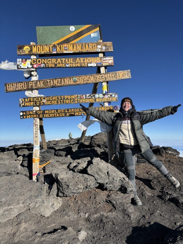 LHOer next to the summit sign of Uhuru Peak on Kilimanjaro at 5,895 meters in great weather with no snow during the Climb Kilimanjaro Expedition with the Life Happens Outdoors team.