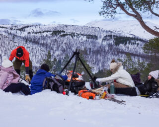 Life Happens Outdoors community members enjoying a winter campfire meal in Senja, Norway, with snowy mountains in the background.