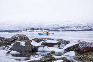 Winter sea kayaking in Senja, Norway with a Life Happens Outdoors guide, paddling across a calm fjord framed by snowy mountains and icy shoreline rocks.