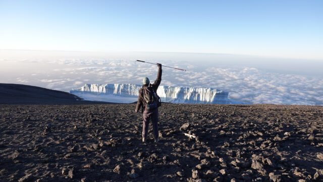 Frederik Sfeir holding his seeing cane above his head with the summit glaciers of Kilimanjaro in front of him and a sea of clouds beneath him at sunrise on the Uhuru Peak summit during the Climb Kilimanjaro Expedition with the Life Happens Outdoors team.