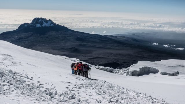 The Life Happens Outdoors team reaching Stella Point as one unit on a snow-covered trail, with the sun blazing, a sea of clouds below, and Mawenzi Crater dominating the skyline in the background during the Climb Kilimanjaro Expedition.