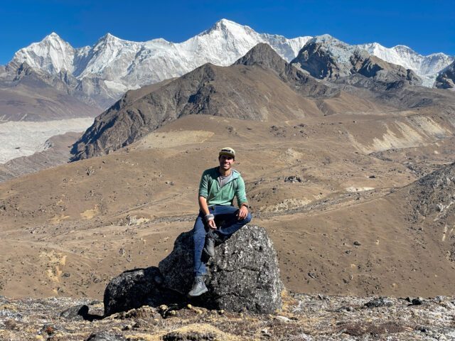 LHO Founder and Team Leader Rami Rasamny sitting on a rock with the South Face of Cho Oyu in the background, in spectacular weather during the Gokyo Lakes Trek with the Life Happens Outdoors team.
