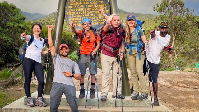 LHOers at the Machame Camp sign at the edge of the rain forest and the short grass vegetation zone on the first day of the Climb Kilimanjaro Expedition on the Machame Route with the Life Happens Outdoors team.