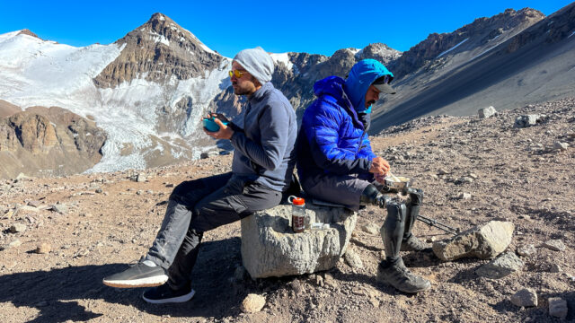 Hari Budha Magar and Frederik Sfeir having breakfast on a rock back to back in Camp 1, Plaza Canadá, against the spectacular backdrop of the Aconcagua glaciers and the Andes in beautiful weather during the Aconcagua Expedition with the Life Happens Outdoors team.
