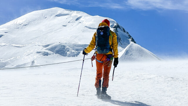 LHO IFMGA guide Pavlos Tsiantos stands atop the Dôme du Goûter, facing Mont Blanc's summit under clear blue skies and brisk winds.