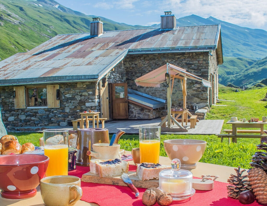 Chalet du Berger with a rustic outdoor breakfast setup featuring local cheeses, bread, and coffee against a backdrop of alpine meadows and mountains.