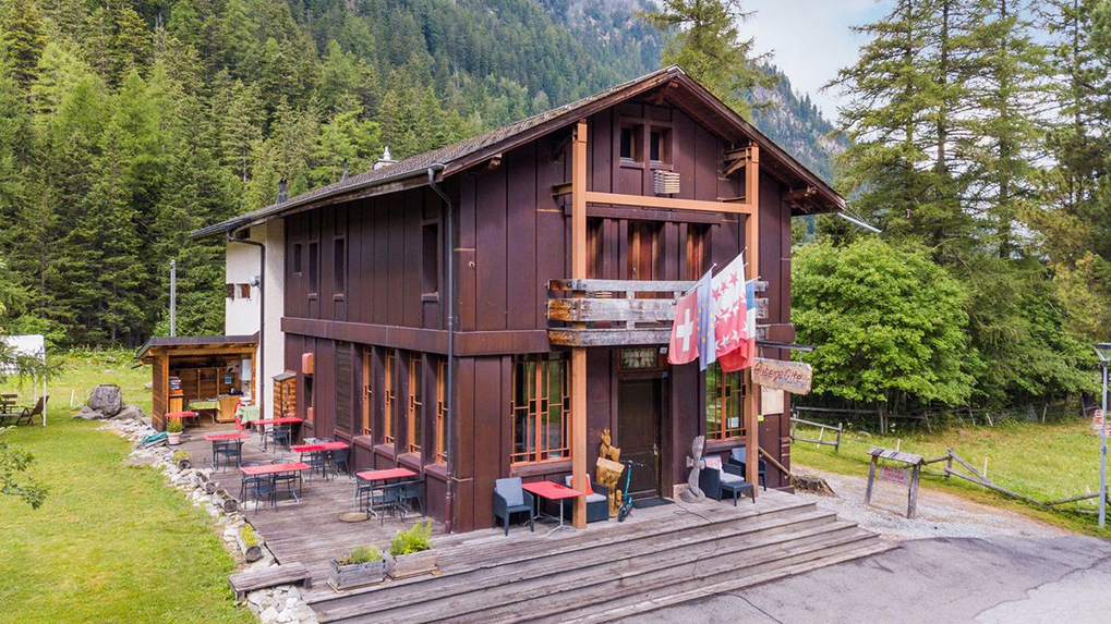 Exterior of Gîte Bon Abri in Champex-Lac, a charming alpine lodge with wooden façade, flower boxes, and views of surrounding forested hills.