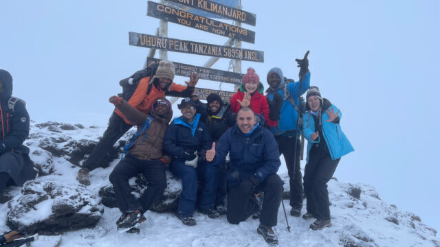 Life Happens Outdoors team of Lamia Farhat, Branca Petrovic, Mohamad Issa, and Hamoud Almutairi standing on Mount Kilimanjaro summit in snowy conditions, celebrating their climb and transformative adventure.