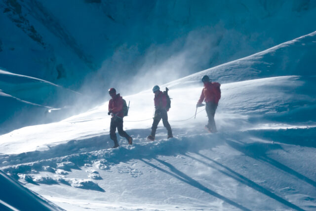Two Life Happens Outdoors climbers with their IFMGA mountain guide approach the Goûter Hut on Mont Blanc in windy alpine conditions during their guided climb.
