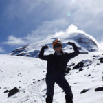 A smiling Life Happens Outdoors climber playfully flexes her biceps in front of Chimborazo under clear, sunny skies, showcasing mountaineering strength and adventure.