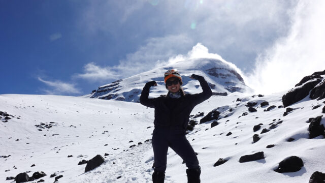 A smiling Life Happens Outdoors climber playfully flexes her biceps in front of Chimborazo under clear, sunny skies, showcasing mountaineering strength and adventure.