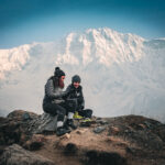 Two Life Happens Outdoors trekkers sit beneath the south face of Annapurna near Annapurna Base Camp after sunset, reflecting on the mountain landscape.