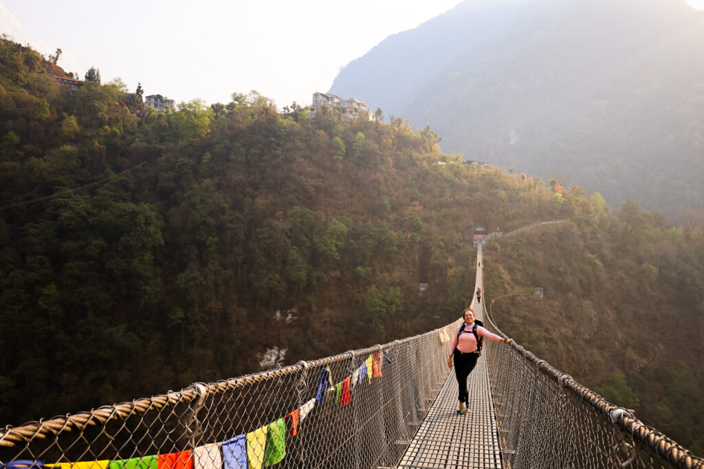 Life Happens Outdoors team leader Ranwa Zahr crossing the new suspension bridge with the village of Jinhu in the background on the Annapurna Base Camp trek.