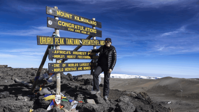 Life Happens Outdoors community member at the Mount Kilimanjaro summit sign with glaciers in the background and a bright blue sky