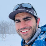 Rami Rasamny smiling in falling snow during a mountain expedition, wearing a blue jacket and cap with snow on his face and clothing, with a winter landscape and bare trees in the background.