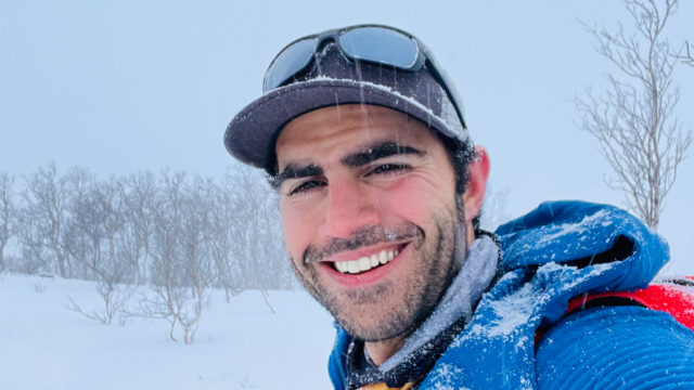 Rami Rasamny smiling in falling snow during a mountain expedition, wearing a blue jacket and cap with snow on his face and clothing, with a winter landscape and bare trees in the background.