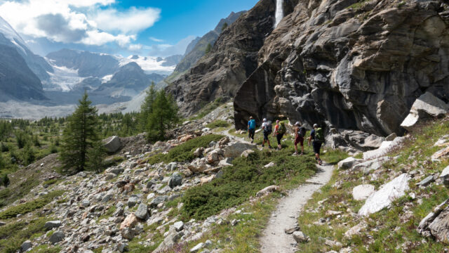 Life Happens Outdoors community members hiking together along an alpine trail beneath a rocky cliff and waterfall with a glacier in the distance
