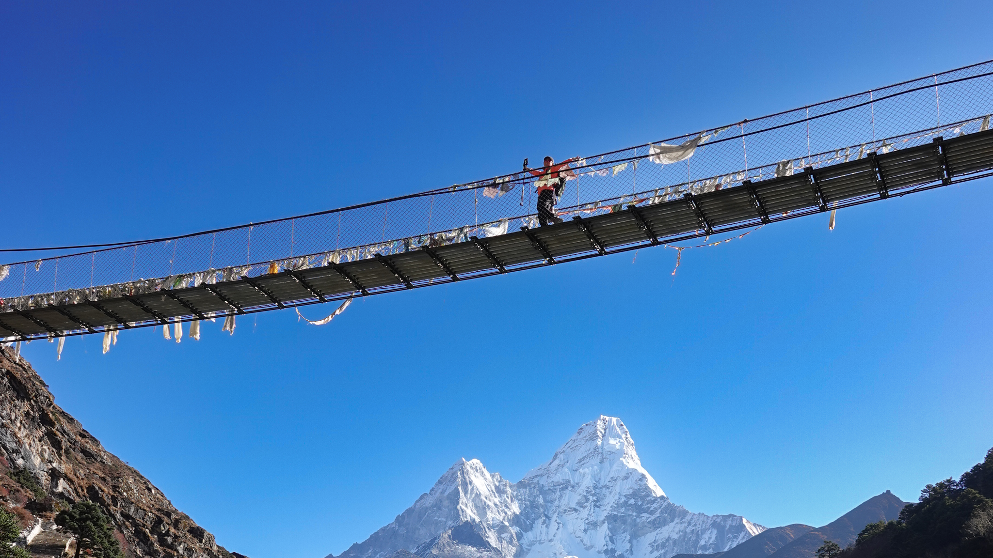 Trekker crossing a suspension bridge in Nepal with a snow covered Himalayan peak behind.