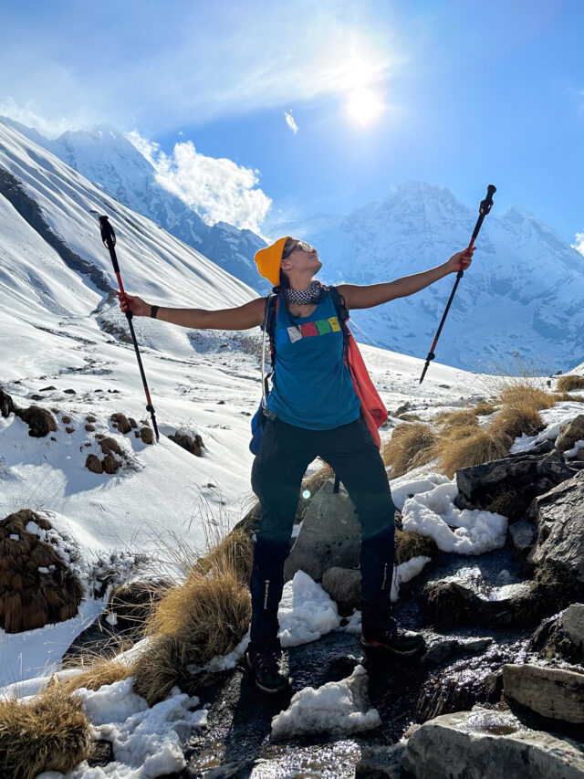 Woman on a snowy trail in Nepal raising trekking poles beneath bright Himalayan peaks