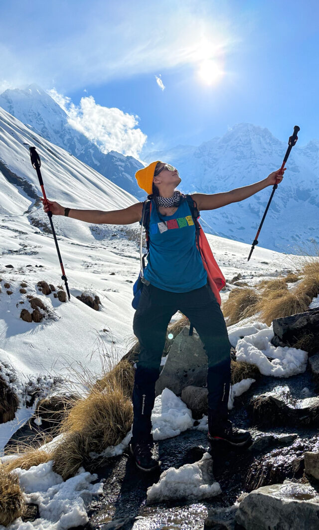 Woman on a snowy trail in Nepal raising trekking poles beneath bright Himalayan peaks