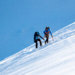 Two climbers ascend a steep snowy mountain slope with ice axes and ropes under a clear blue sky.