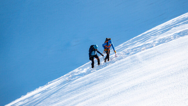 Two climbers ascend a steep snowy mountain slope with ice axes and ropes under a clear blue sky.