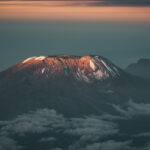 Sunrise light on Kilimanjaro summit plateau with snow patches, dark volcanic slopes and Mawenzi rising in the background above clouds.