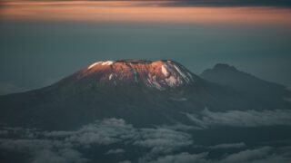 Sunrise light on Kilimanjaro summit plateau with snow patches, dark volcanic slopes and Mawenzi rising in the background above clouds.