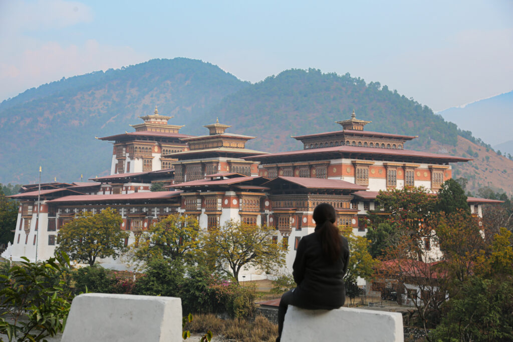 A traveler looks out toward a traditional Himalayan monastery fortress framed by forested mountains, reflecting on the journey and shared adventure