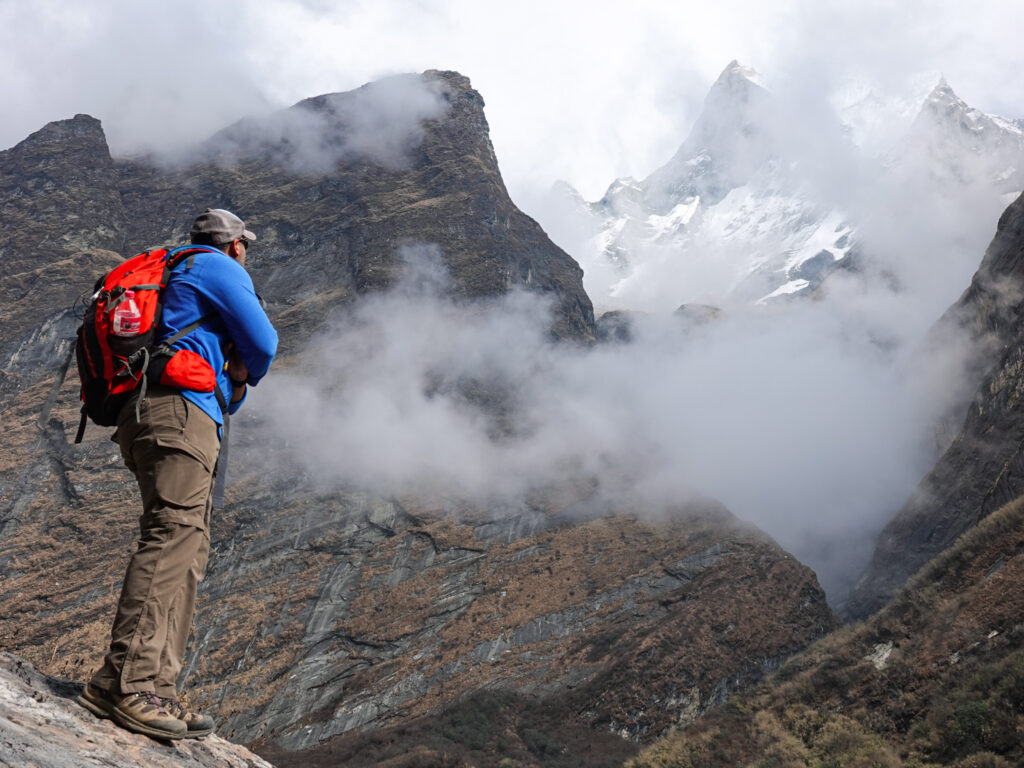 A trekker with a red backpack pauses to look toward cloud wrapped mountain peaks on a dramatic alpine trail.