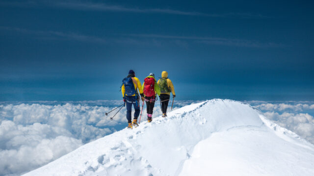 Three climbers walking the Mont Blanc summit ridge above a sea of clouds.