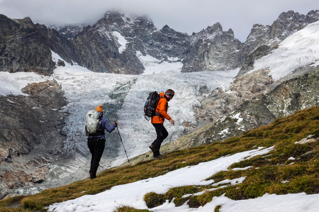 Two trekkers navigating snow and mixed terrain on the Grand Col Ferret approach to the Tour du Mont Blanc in early season conditions