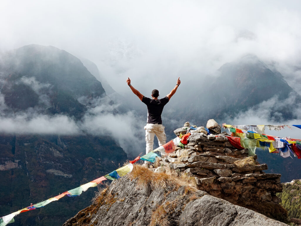 Trekker standing above the clouds in Nepal during an Everest Base Camp trek with prayer flags and Himalayan mountains in the background