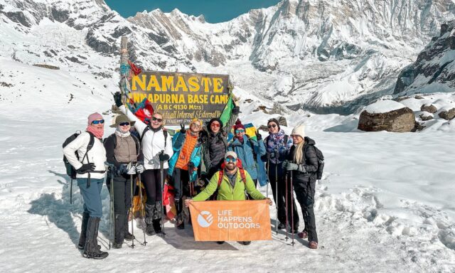 Life Happens Outdoors trekkers stand by the Annapurna Base Camp sign beneath snowy peaks.