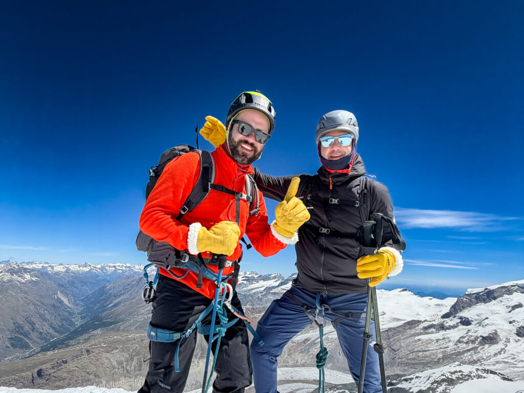 Two climbers in helmets and harnesses smiling on a snowy alpine mountain during a high altitude preparation climb with Life Happens Outdoors.