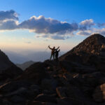 Three Life Happens Outdoors adventurers standing together on a rocky mountain ridge at sunrise.