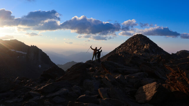 Three Life Happens Outdoors adventurers standing together on a rocky mountain ridge at sunrise.