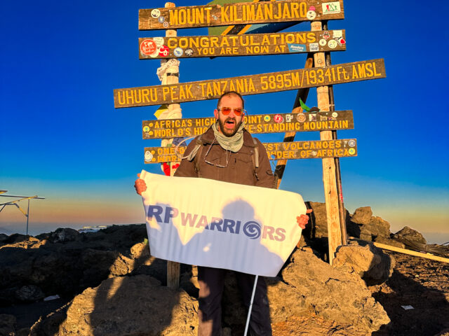 Fred Sfeir at the Uhuru Peak sign on Kilimanjaro holding an RP Warriors banner during the Moving Mountains documentary.