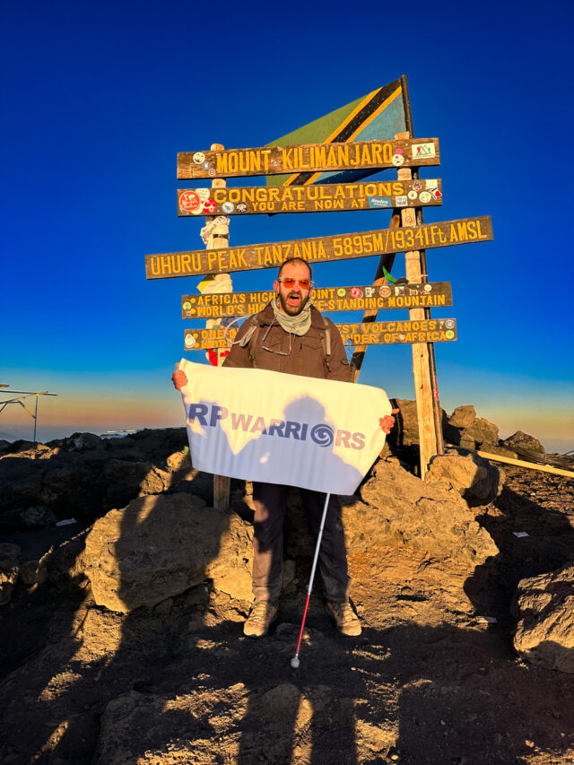 Fred Sfeir at the Uhuru Peak sign on Kilimanjaro holding an RP Warriors banner during the Moving Mountains documentary.
