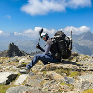 Trekker resting with a large backpack on a rocky mountain ridge beneath a blue sky.