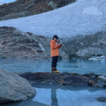 Trekker in an orange jacket holding an open notebook beside a glacial lake below a mountain glacier.