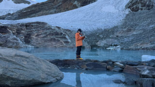 Trekker in an orange jacket holding an open notebook beside a glacial lake below a mountain glacier.