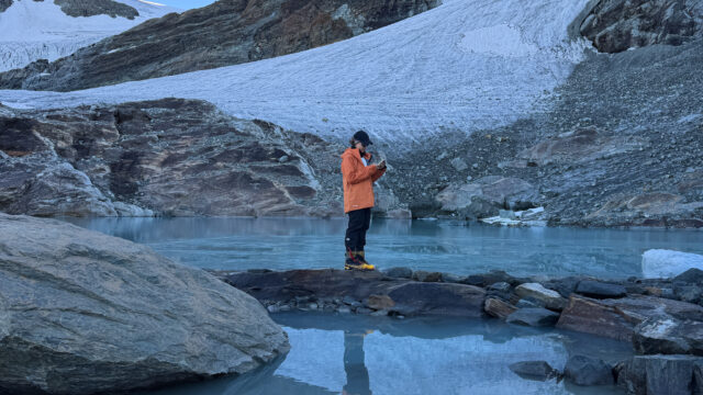 Trekker in an orange jacket holding an open notebook beside a glacial lake below a mountain glacier.
