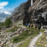 Trekkers following a rocky alpine trail beneath a waterfall with a glacier across the valley