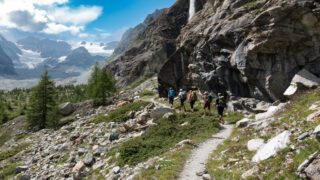 Trekkers following a rocky alpine trail beneath a waterfall with a glacier across the valley