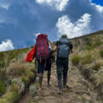 Two trekkers carrying backpacks ascend a rocky high-altitude mountain trail.