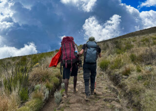 Two trekkers carrying backpacks ascend a rocky high-altitude mountain trail.