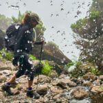 Life Happens Outdoors trekker walking across rocky terrain beside a mountain waterfall.