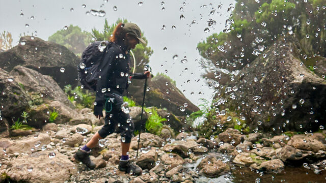 Life Happens Outdoors trekker walking across rocky terrain beside a mountain waterfall.