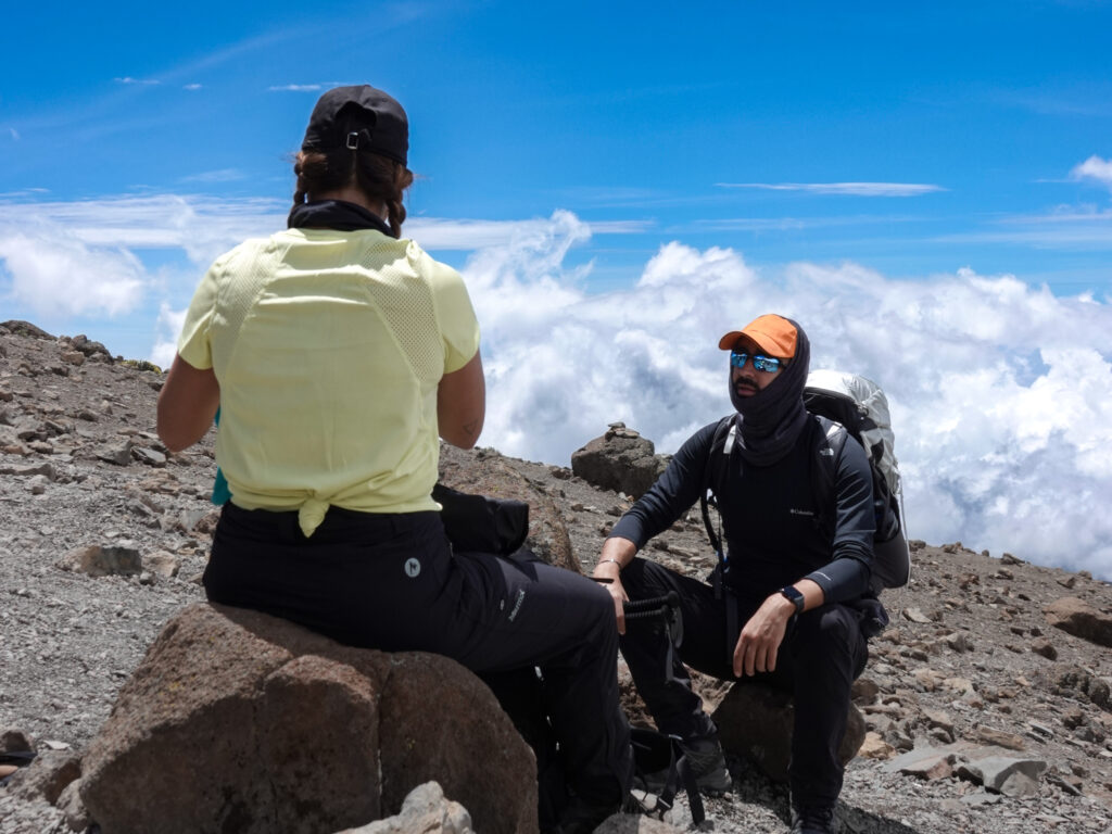 Two Life Happens Outdoors trekkers resting on a rocky high altitude slope above the clouds.